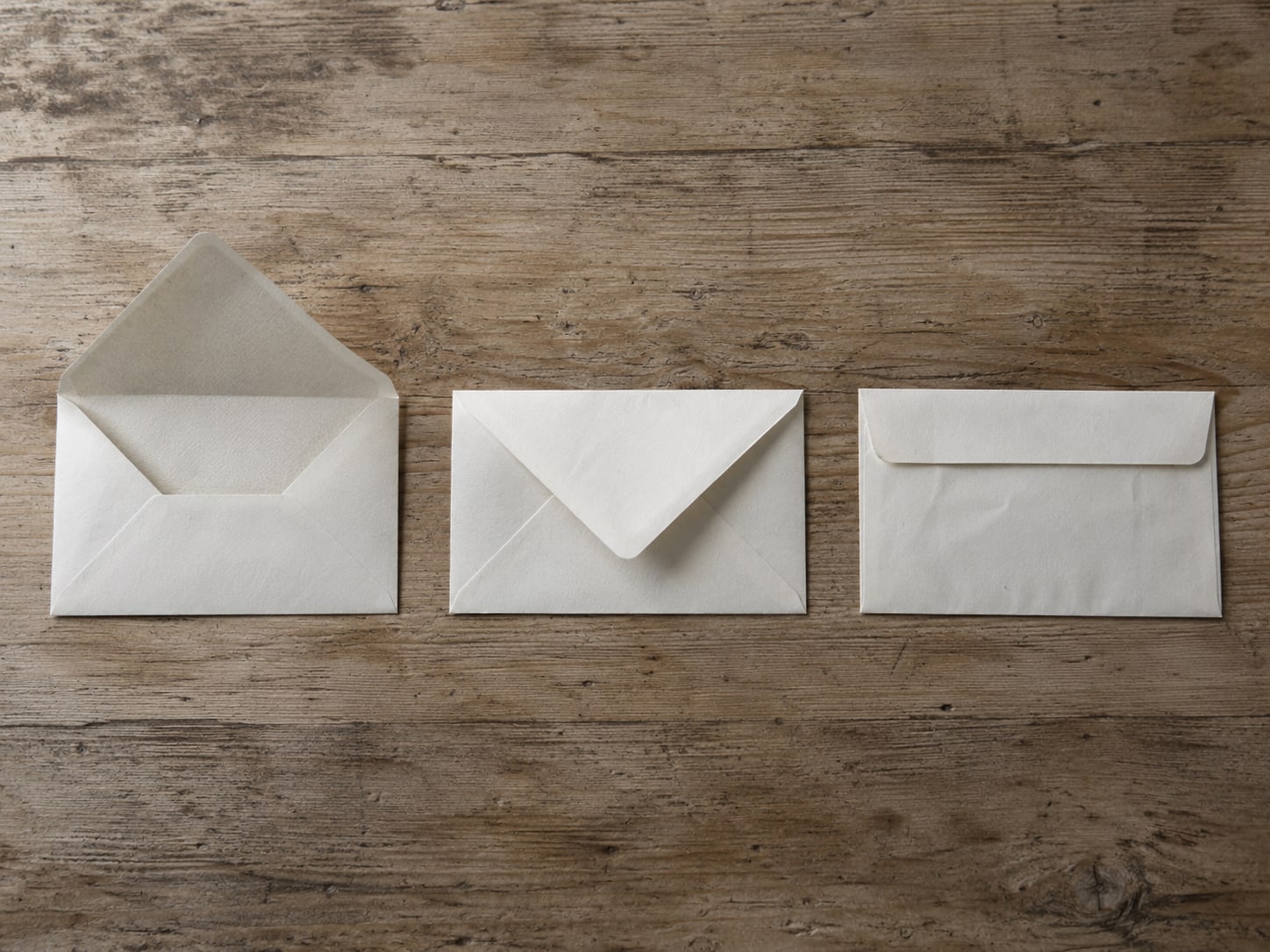 A set of three plain paper envelopes in a row on a wooden counter, one open, one sealed and one with its flap tucked in.