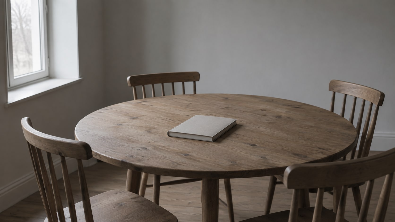 A round wooden table with several empty chairs pulled up around it and a blank notebook in the middle.