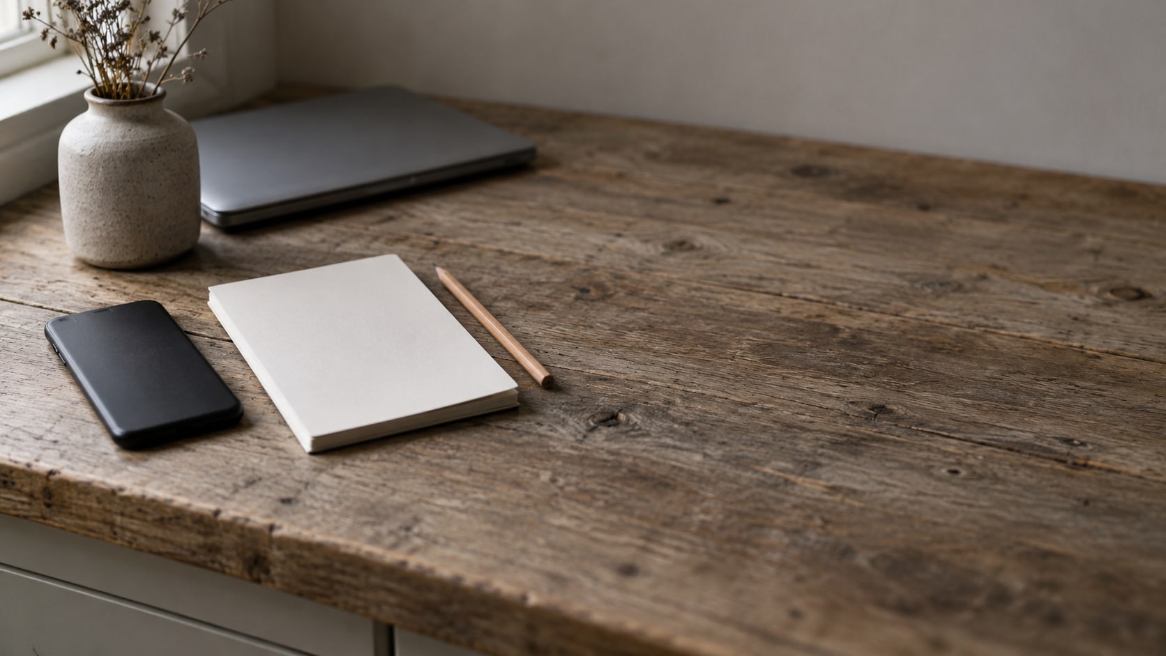 A closed smartphone lying face down beside a blank notepad and a pencil on a wooden counter.