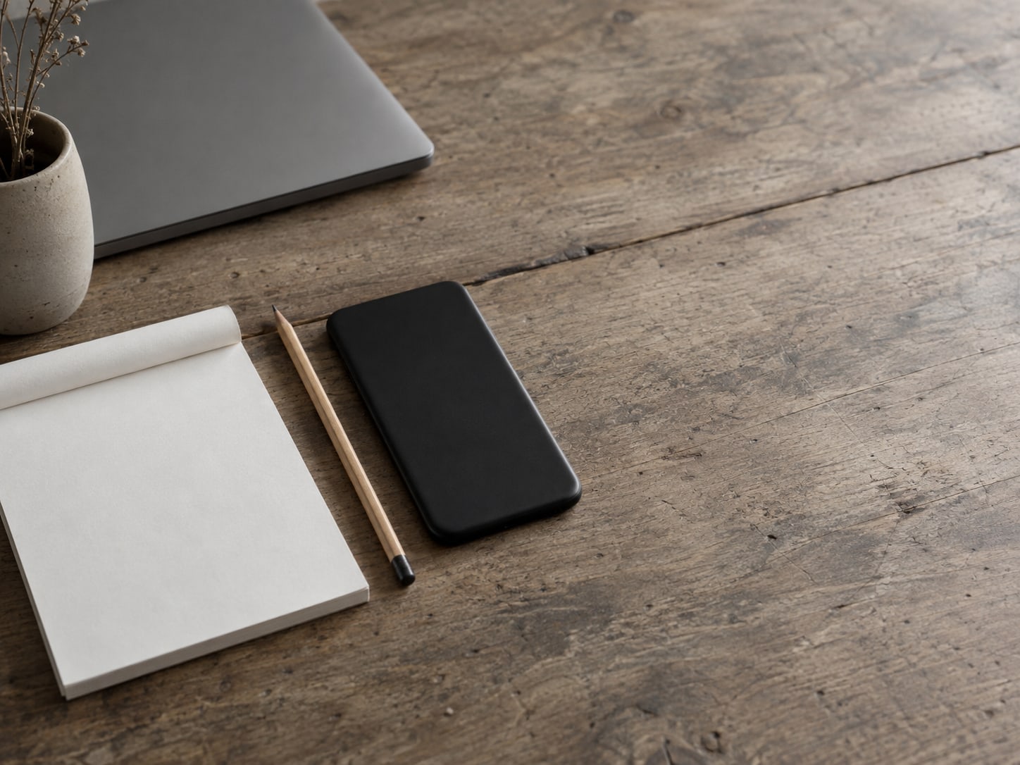 A closed phone lying face down beside a pencil and a blank notepad on a plain wooden counter.