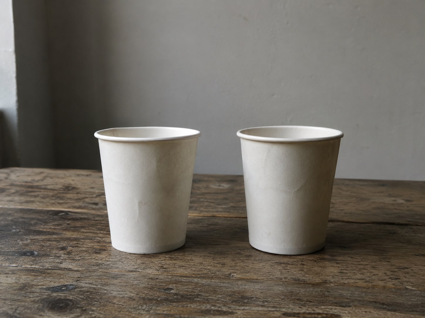 A pair of identical blank paper cups on a wooden table, one slightly apart from the other.