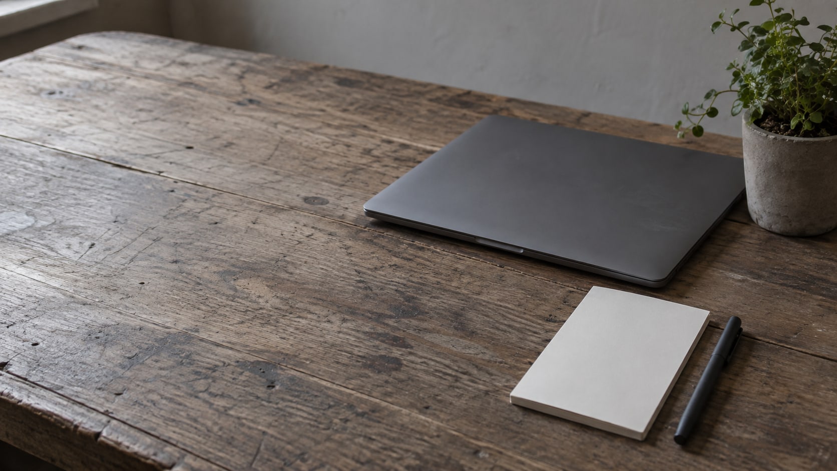 A closed laptop, a blank notepad and a pen on a worn wooden desk beside a small potted plant.