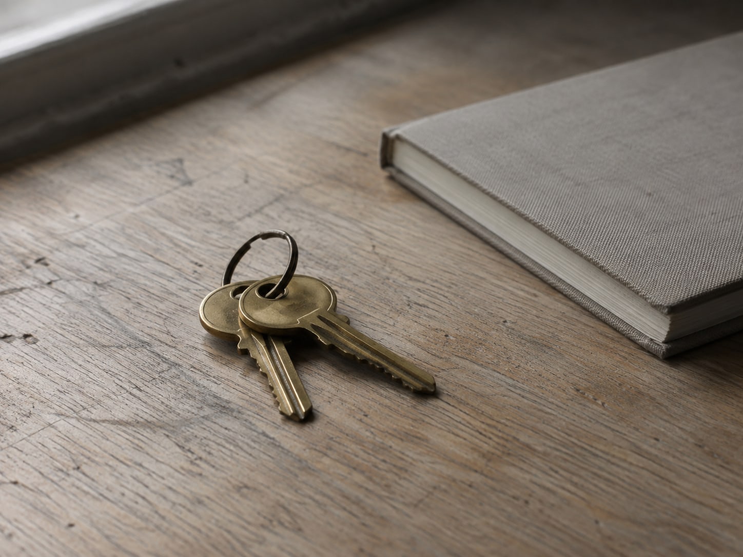 Two brass keys on a plain ring resting on a wooden desk next to a closed notebook.