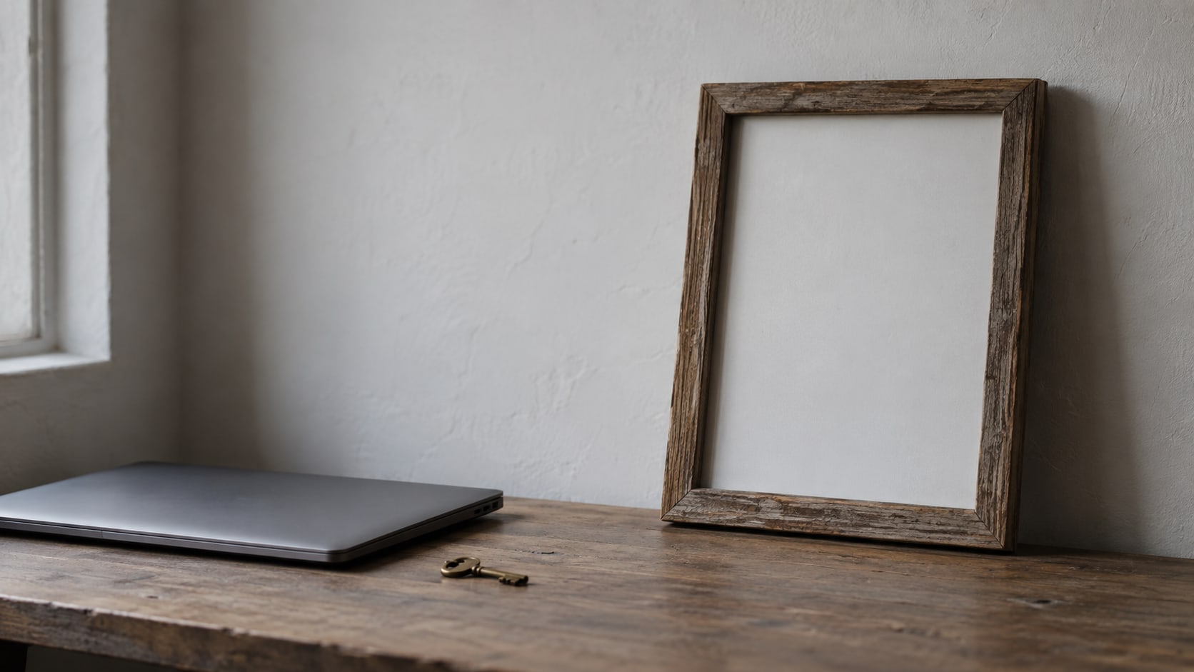 An empty wooden picture frame leaning against a wall on a desk beside a closed laptop and a small brass key.
