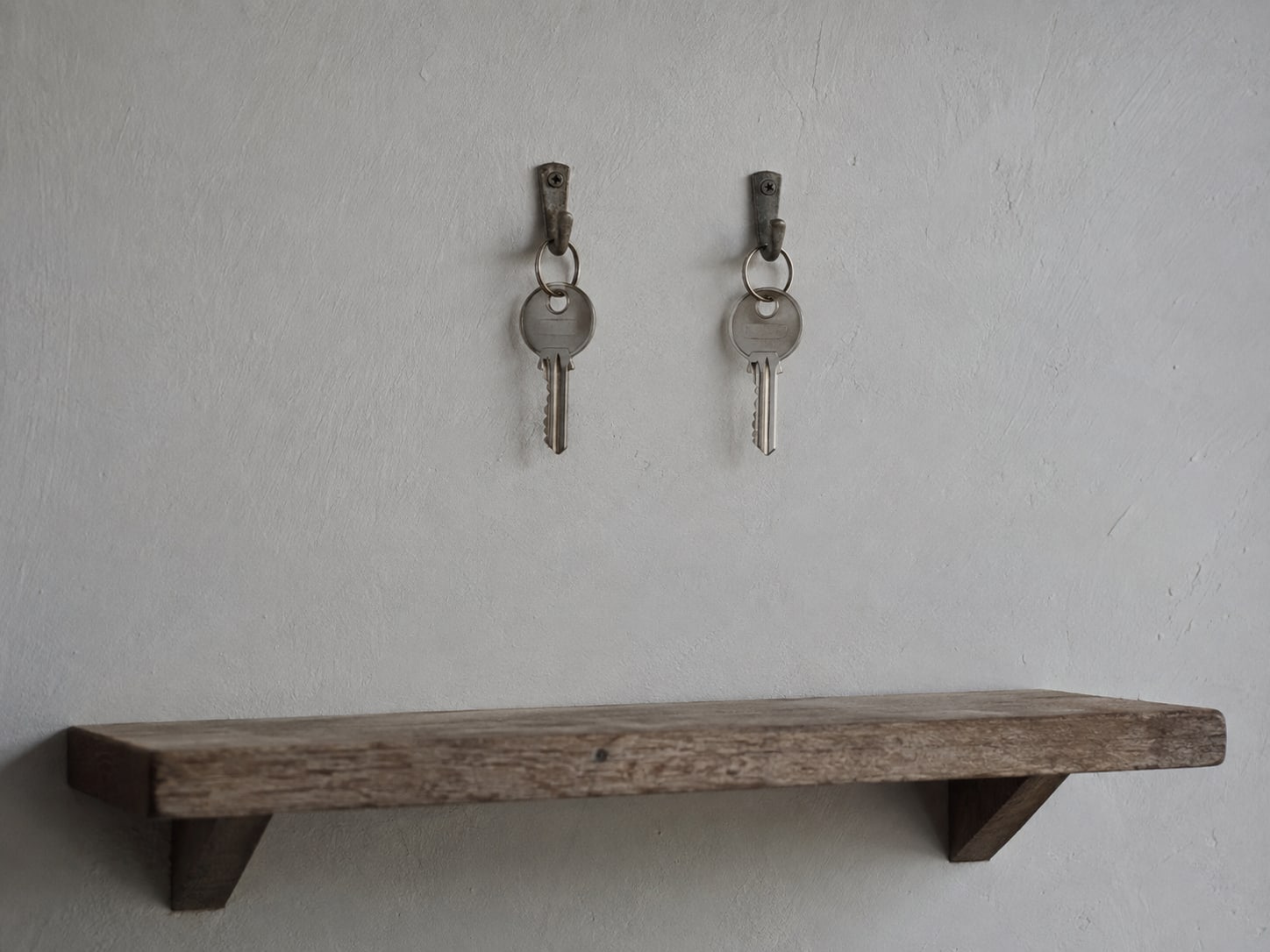 Two identical house keys hanging on separate hooks on a plain wall above a small wooden shelf.