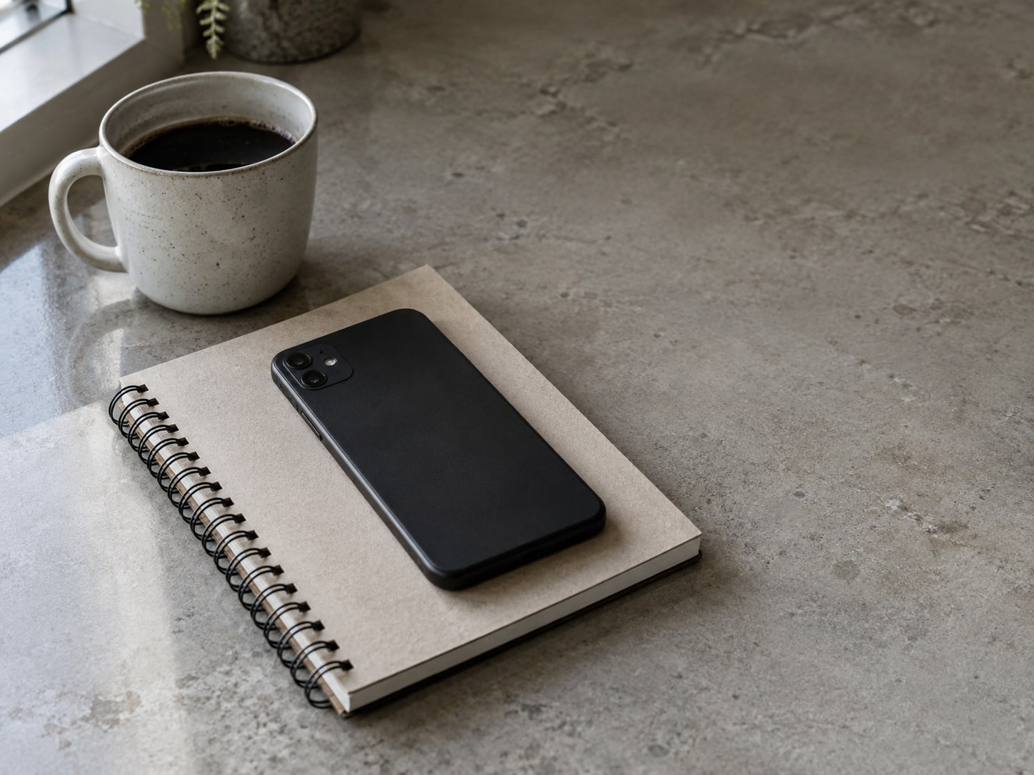 A face-down phone resting on a closed spiral notebook beside a cooling cup of coffee on a kitchen counter.