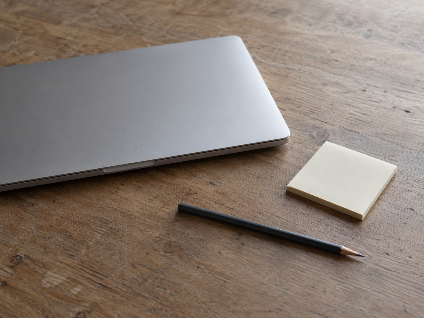 A closed laptop on a wooden desk beside a small pad of blank sticky notes and a pen.
