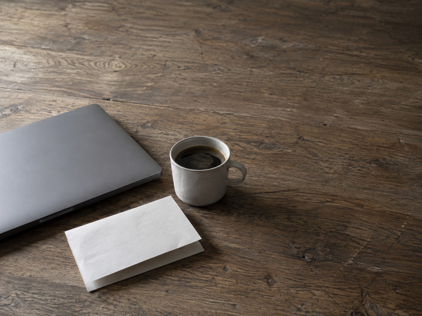 A closed laptop beside a single folded letter and a cup of coffee on a plain wooden desk.
