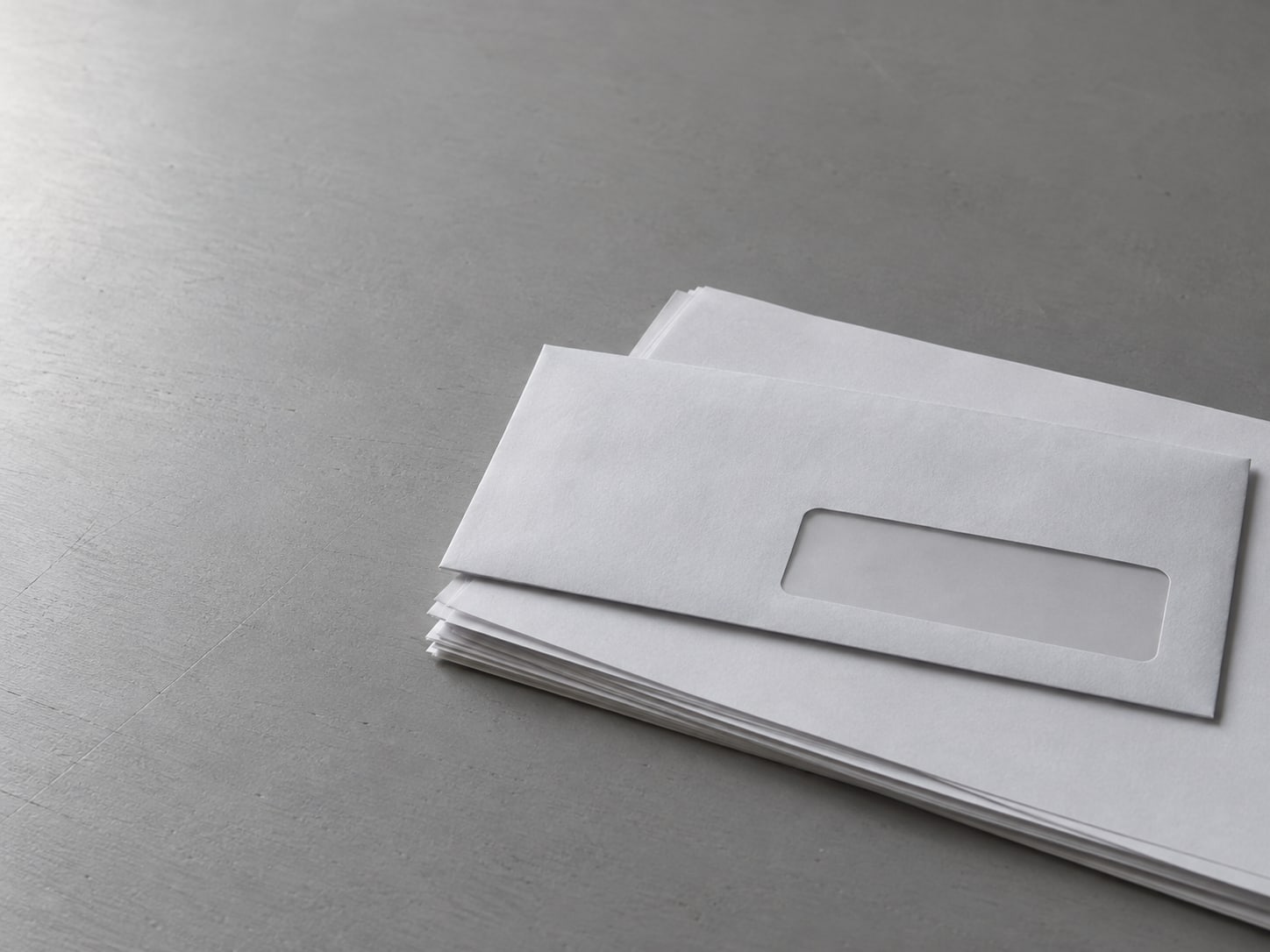 An unopened window envelope resting on a small stack of blank papers on a gray desk.