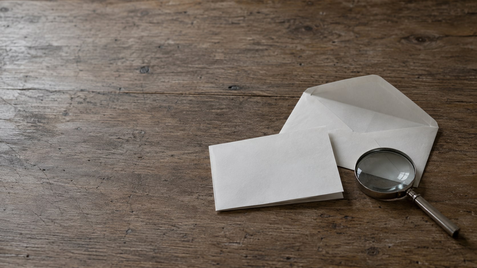 An opened plain envelope and a folded blank sheet of paper beside a magnifying glass on a wooden desk.