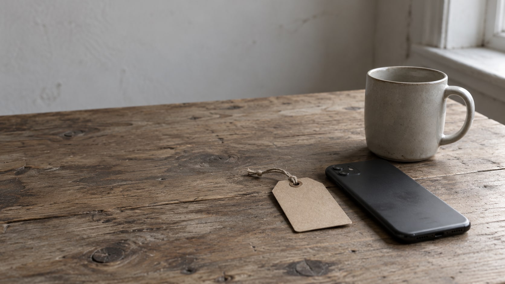 A phone lying face down beside a blank cardboard tag and a ceramic mug on a worn wooden desk.