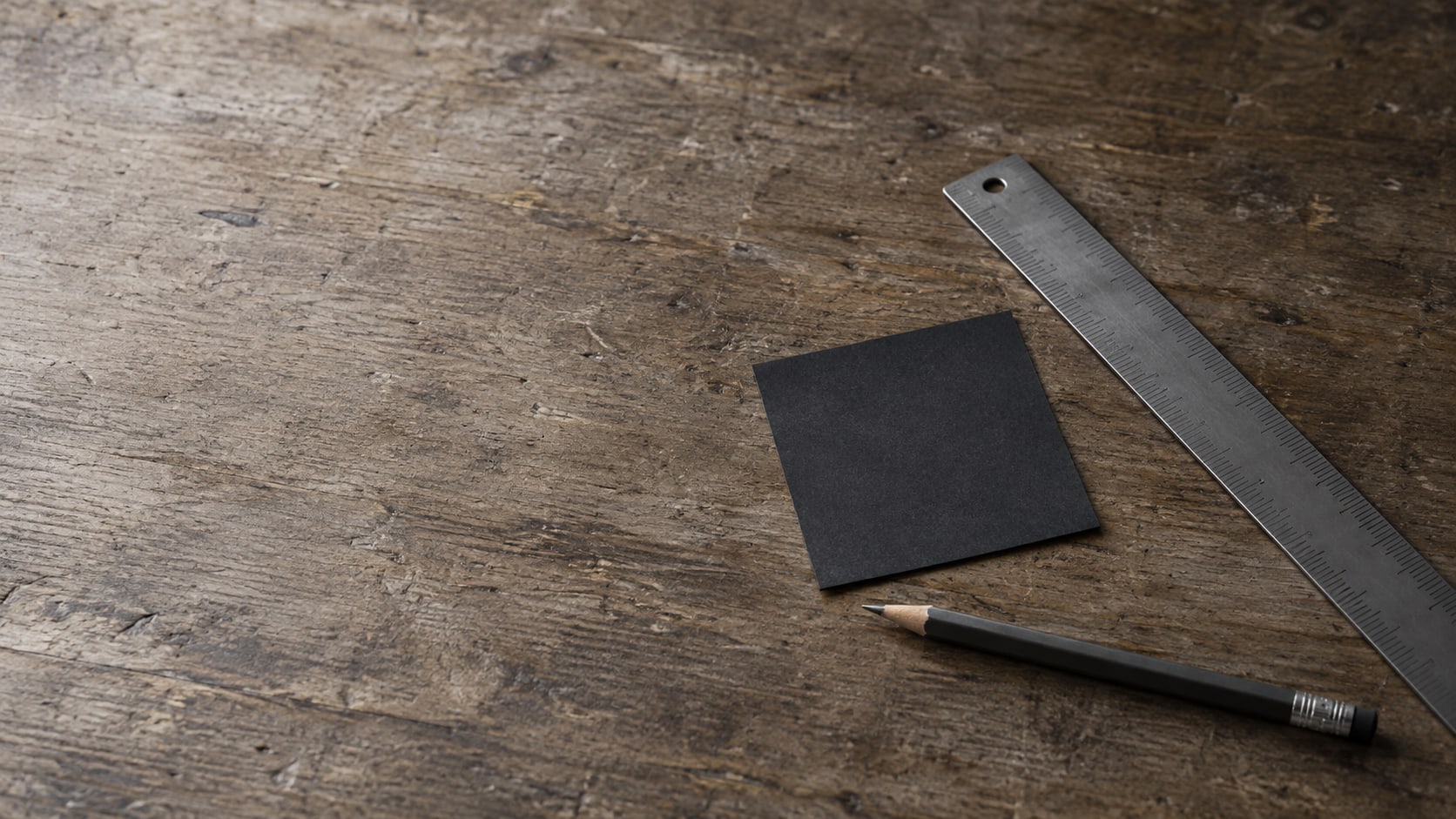 A small square of dark paper beside a ruler and a pencil on a worn wooden desk.