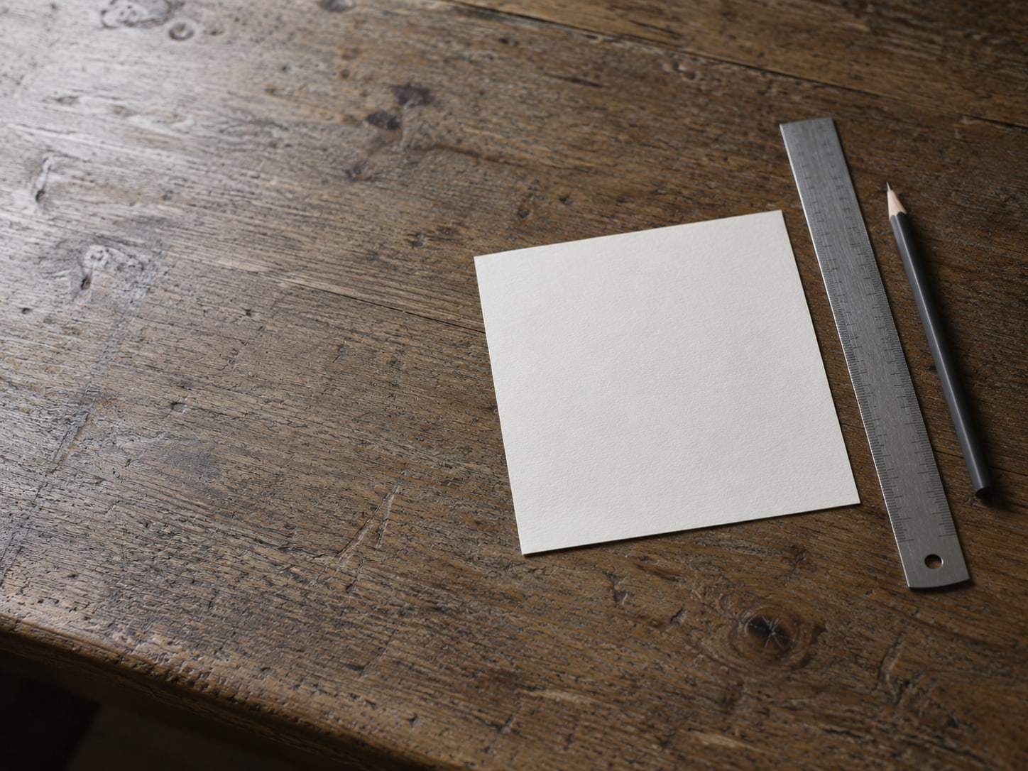 A single sheet of blank paper and a ruler lying square on a worn wooden desk beside a pencil.
