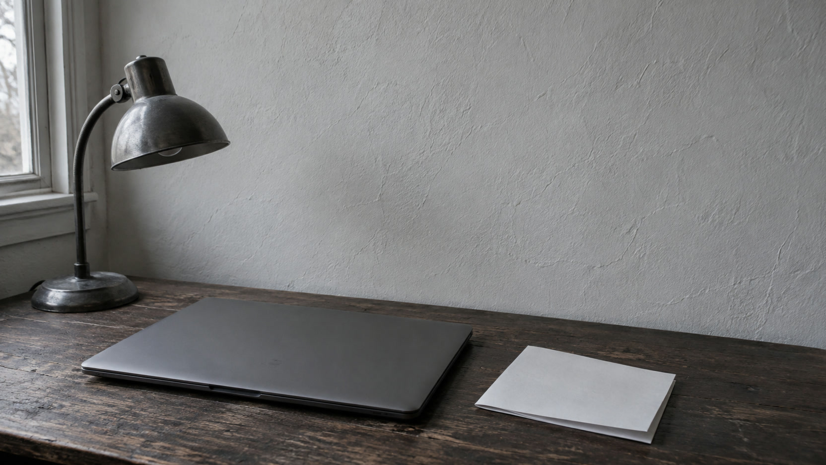 A closed laptop with a small desk lamp switched off beside it and a folded sheet of plain paper on a dark wooden desk.