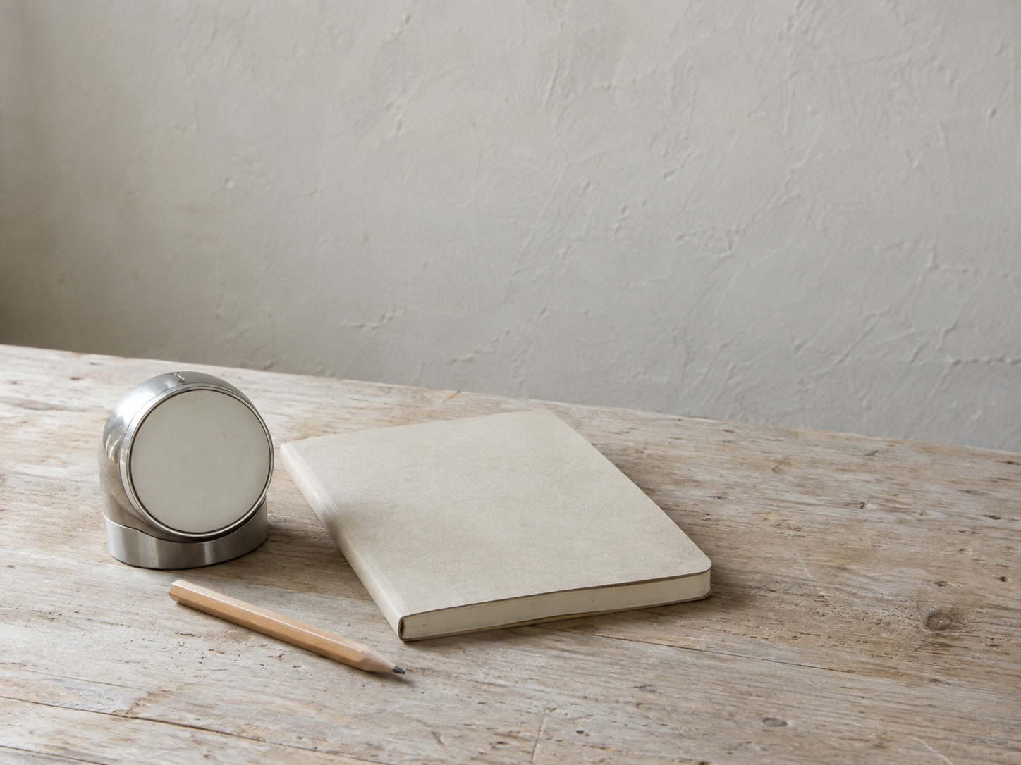 A small kitchen timer beside a closed notebook and a short pencil on a light wooden table.