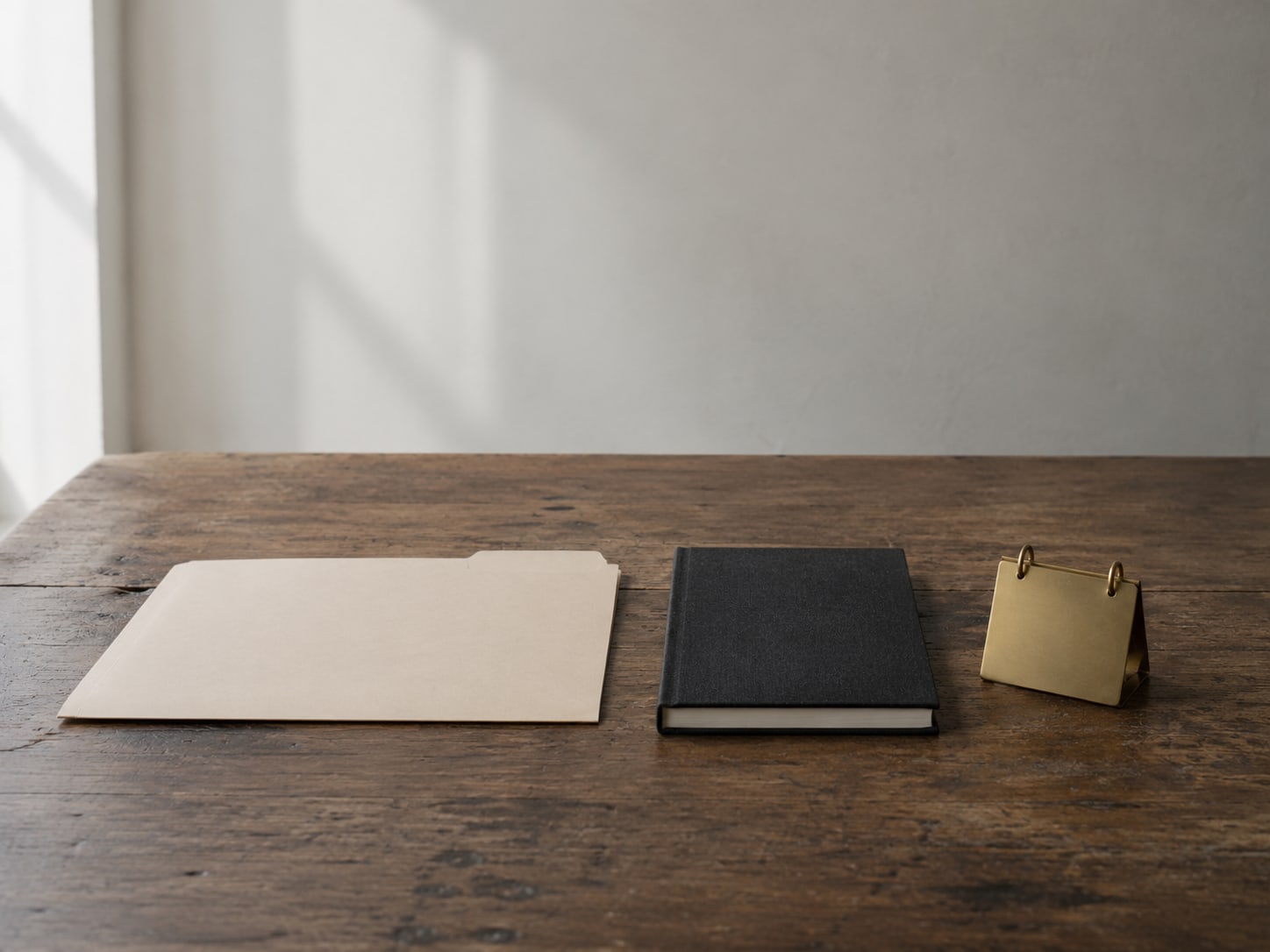 A manila folder, a closed appointment book and a small brass desk calendar lined up on a wooden table.