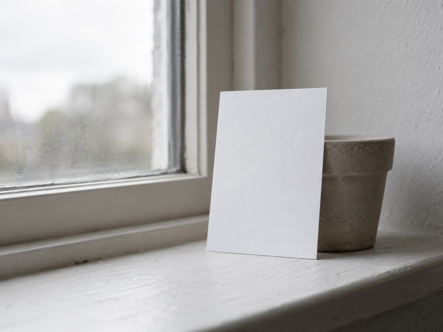 A single printed card with a blank white face, propped upright against a small plant pot on a windowsill.