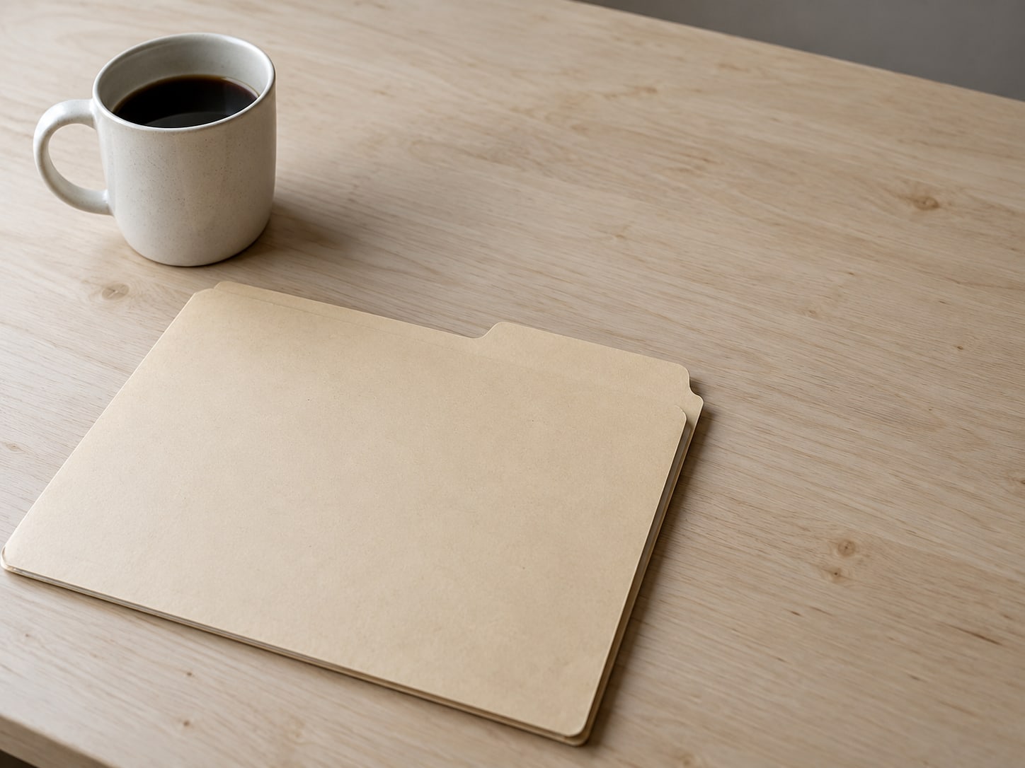 A closed manila folder with a blank tab on a clean desk beside a ceramic coffee mug.