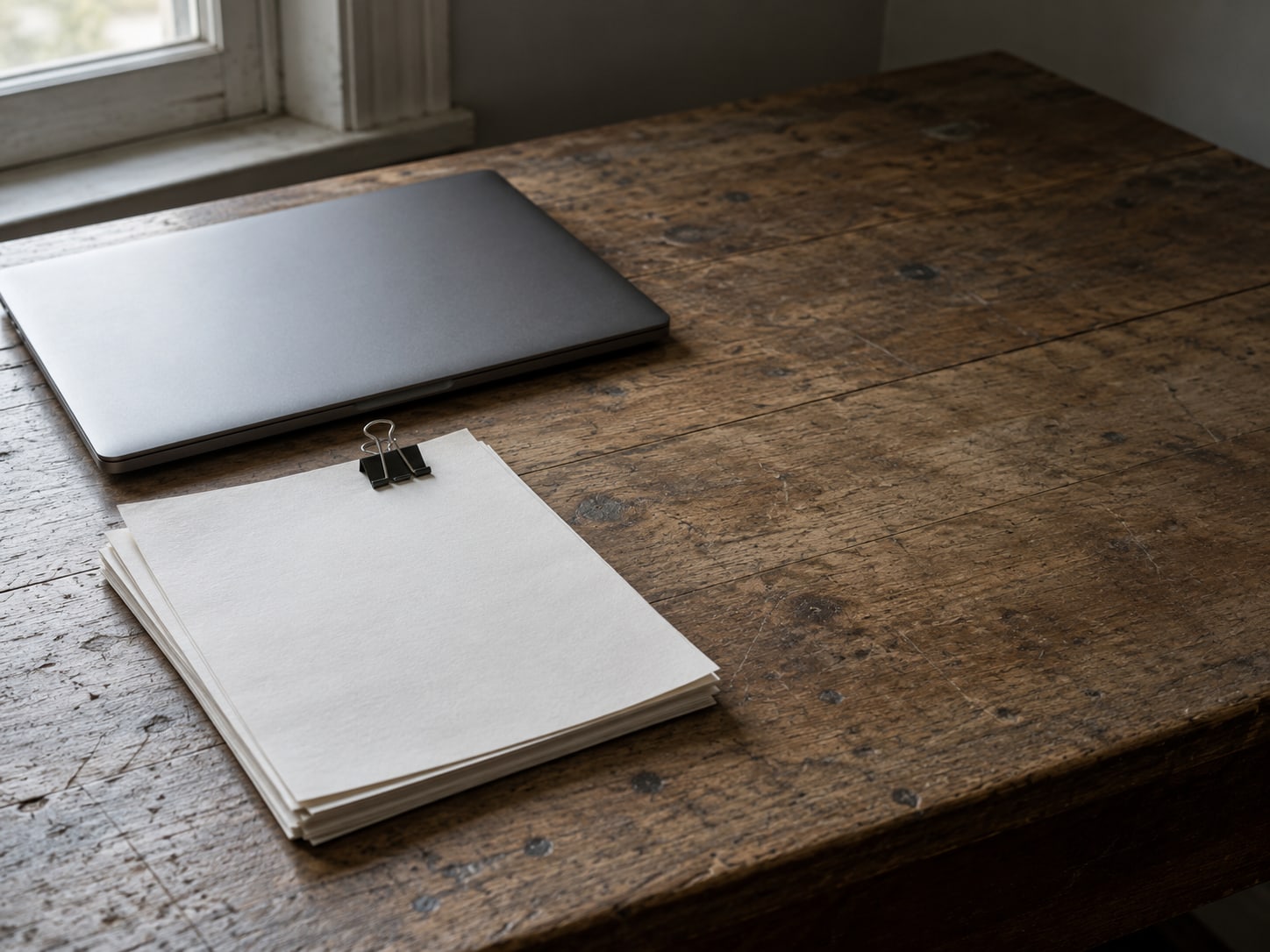 A closed laptop beside a small stack of blank paper estimates held together with a binder clip on a worn wooden desk.