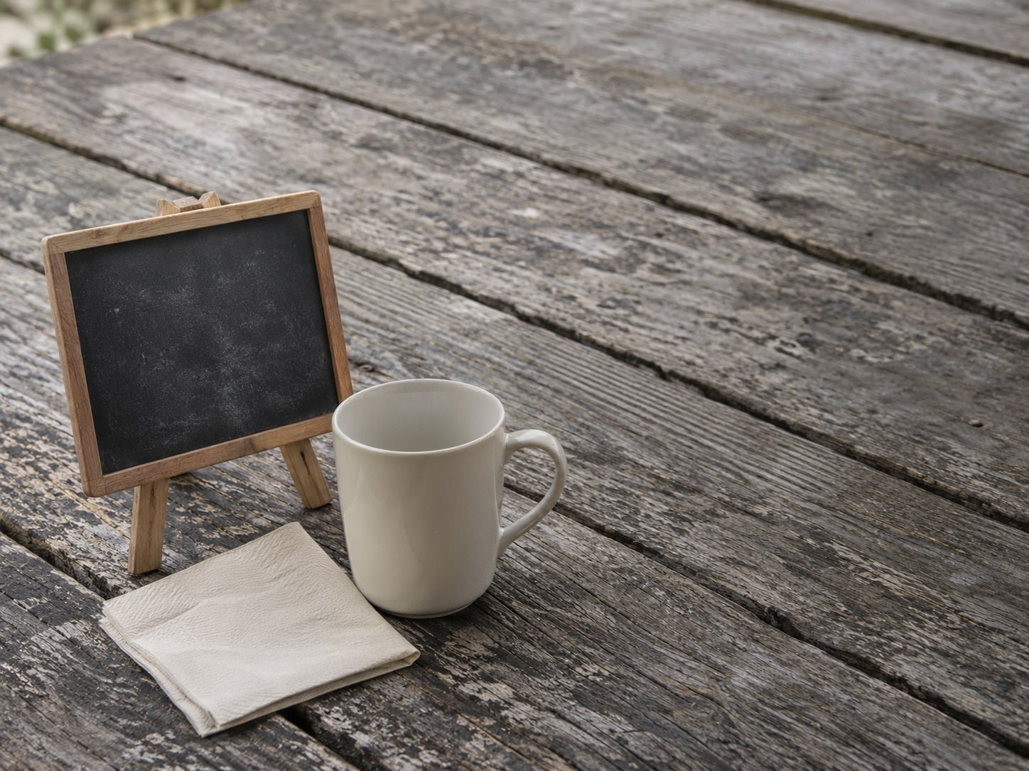 Una pizarra de color negro en blanco sobre un pequeño caballete, junto a una servilleta de papel doblada y una taza de café vacía sobre una mesa de picnic.