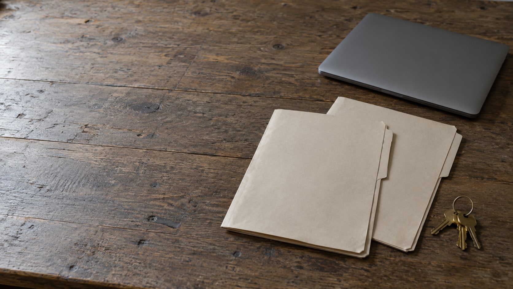 A closed laptop and two blank folders side by side on a wooden desk beside a ring of brass keys.