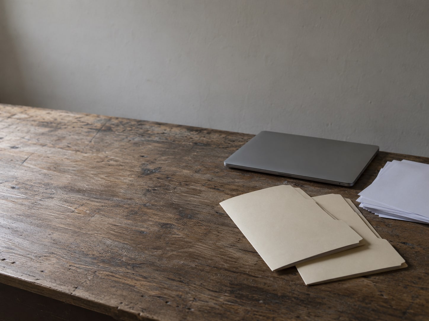 A closed laptop and a small stack of blank paper beside two folders on a wooden desk.