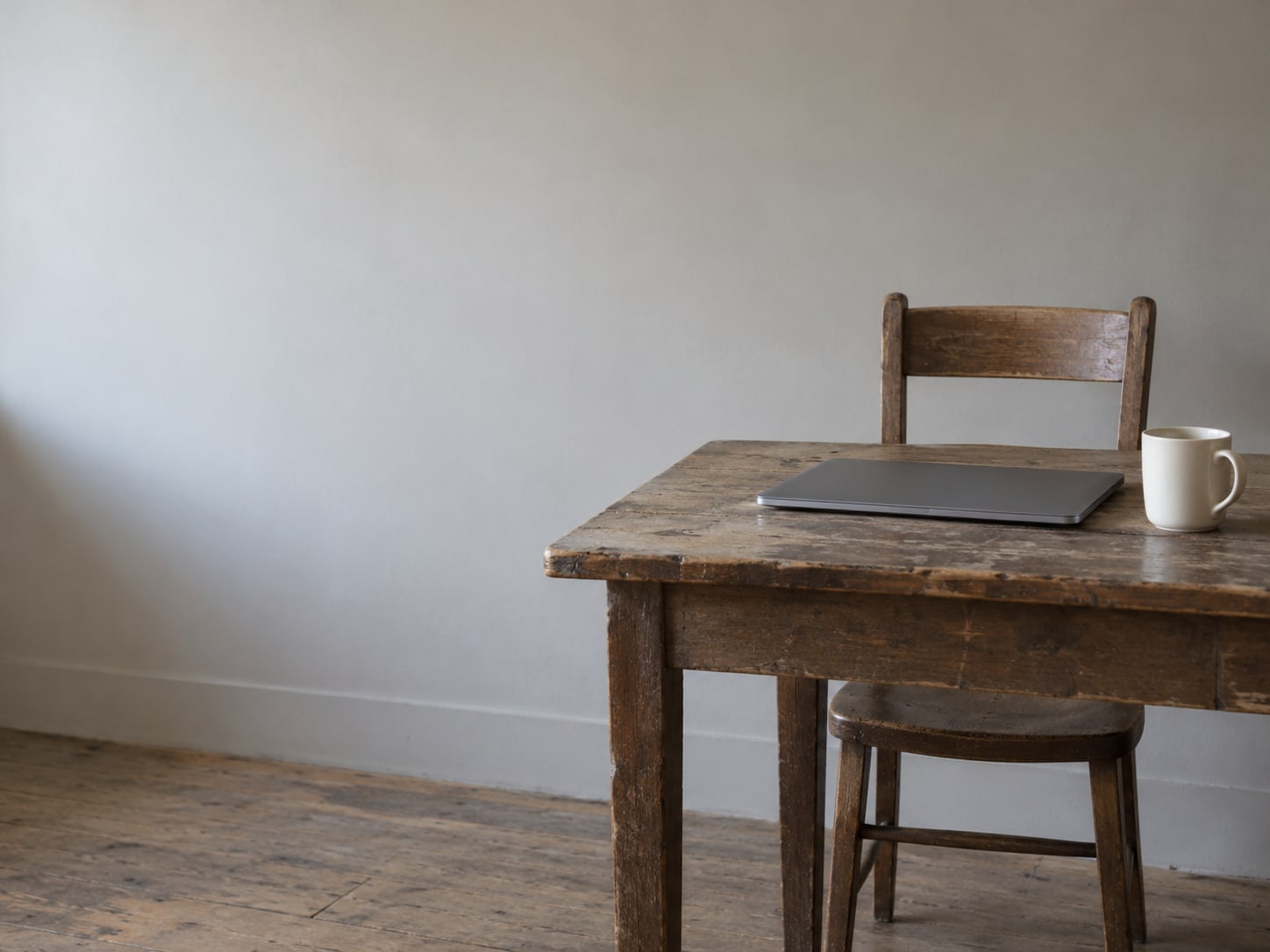 An empty chair pulled up to a desk with a closed laptop and a ceramic mug.