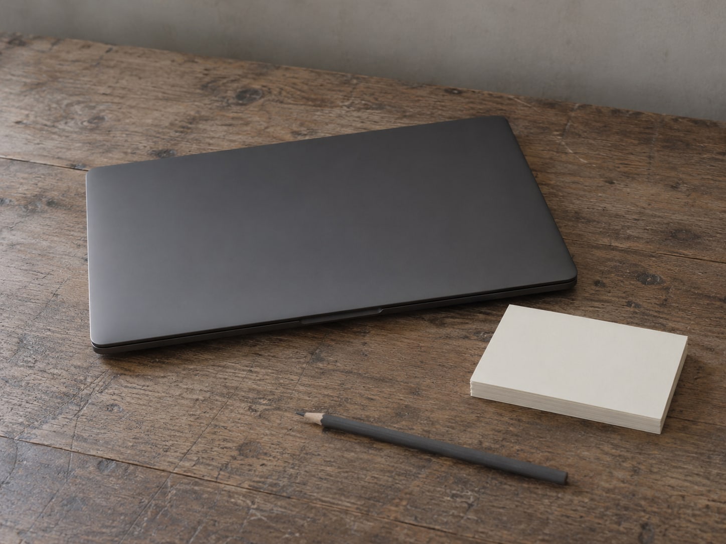 Una laptop cerrada junto a una pequeña pila de fichas en blanco y un lápiz sobre un escritorio de madera gastado.