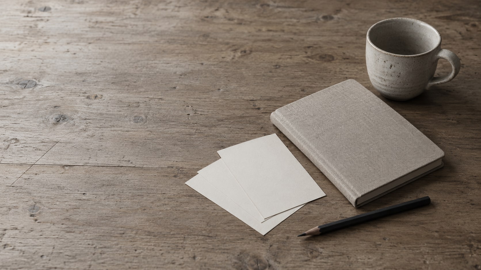 A closed notebook, a pencil and three blank paper slips fanned out on a wooden desk beside a small empty coffee cup.
