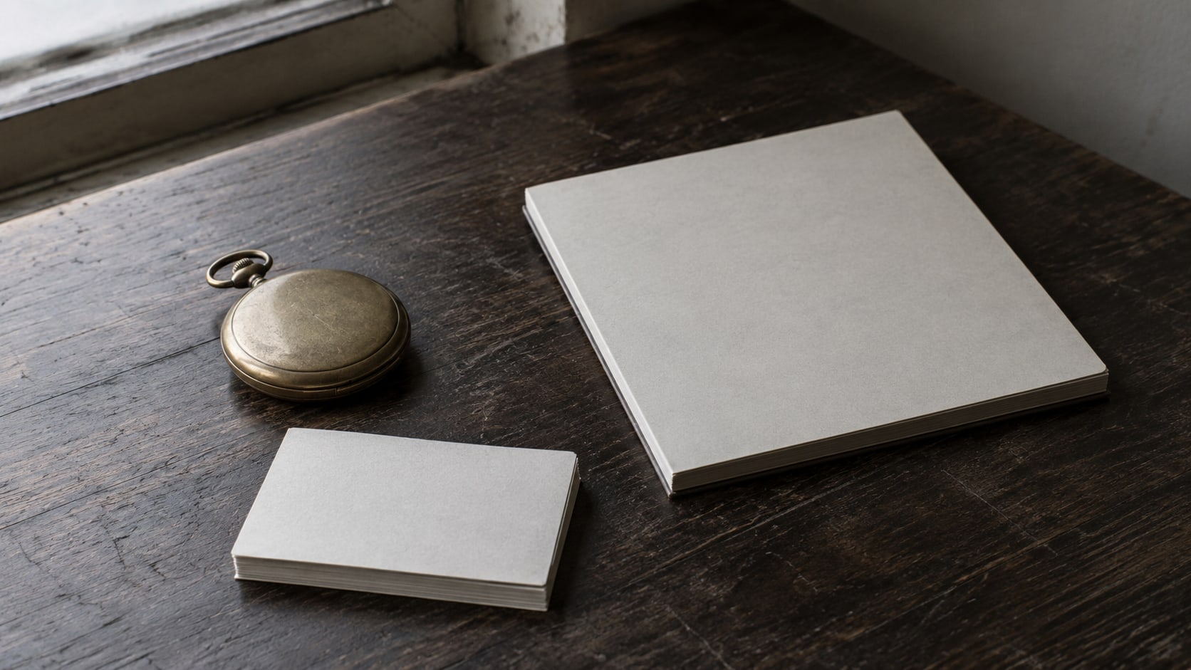 A brass pocket watch and a closed paper calendar on a dark wooden table beside a small stack of blank index cards.