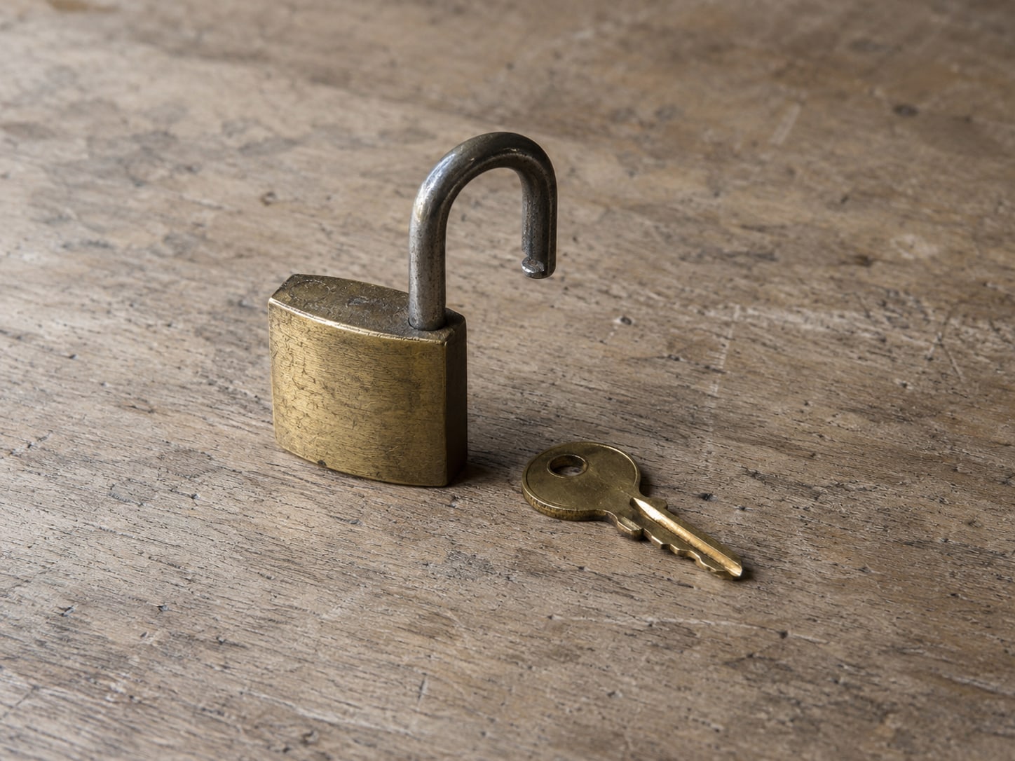A single unlocked padlock resting on a flat wooden board next to a small brass key.
