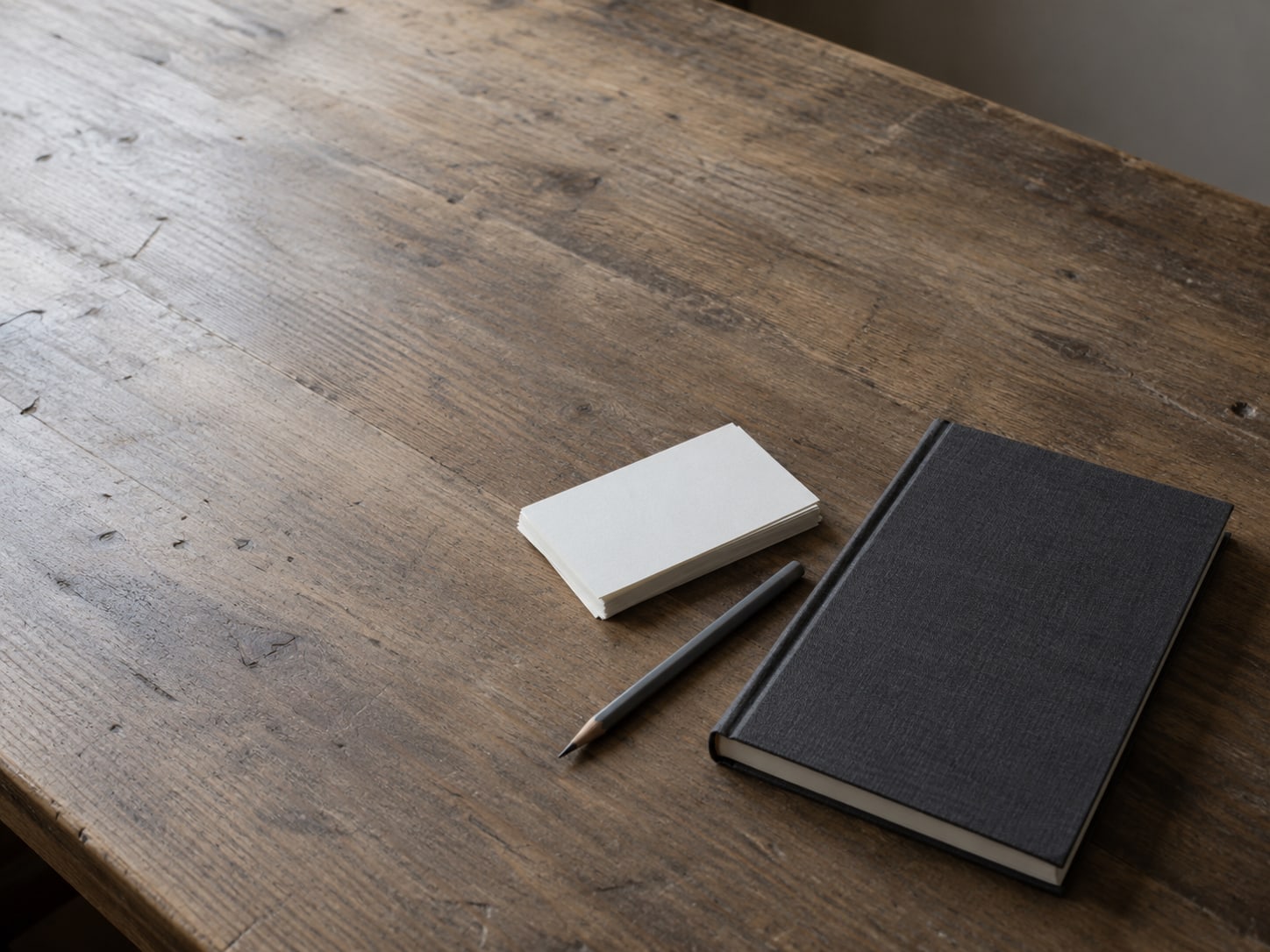 Una libreta cerrada y un lápiz sobre un escritorio de madera, junto a un pequeño montón de fichas en blanco.