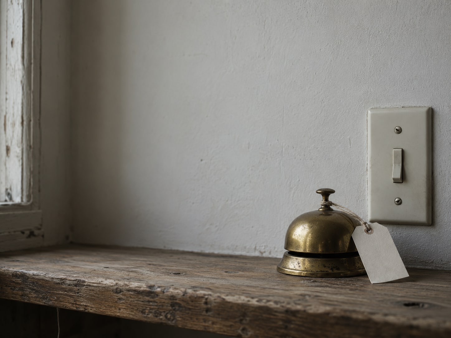 A small wall light switch plate beside a brass desk bell, both on a wooden shelf, with a blank paper tag tied to the bell.