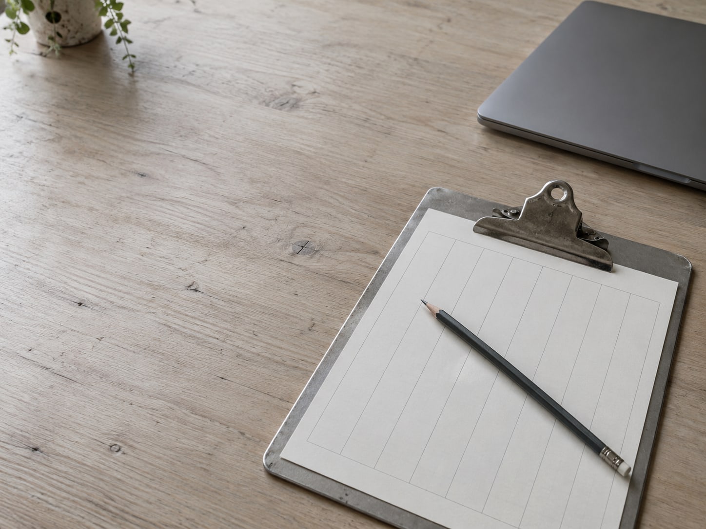 A pencil lying across a blank printed grid of seven columns on a clipboard, next to a closed laptop.