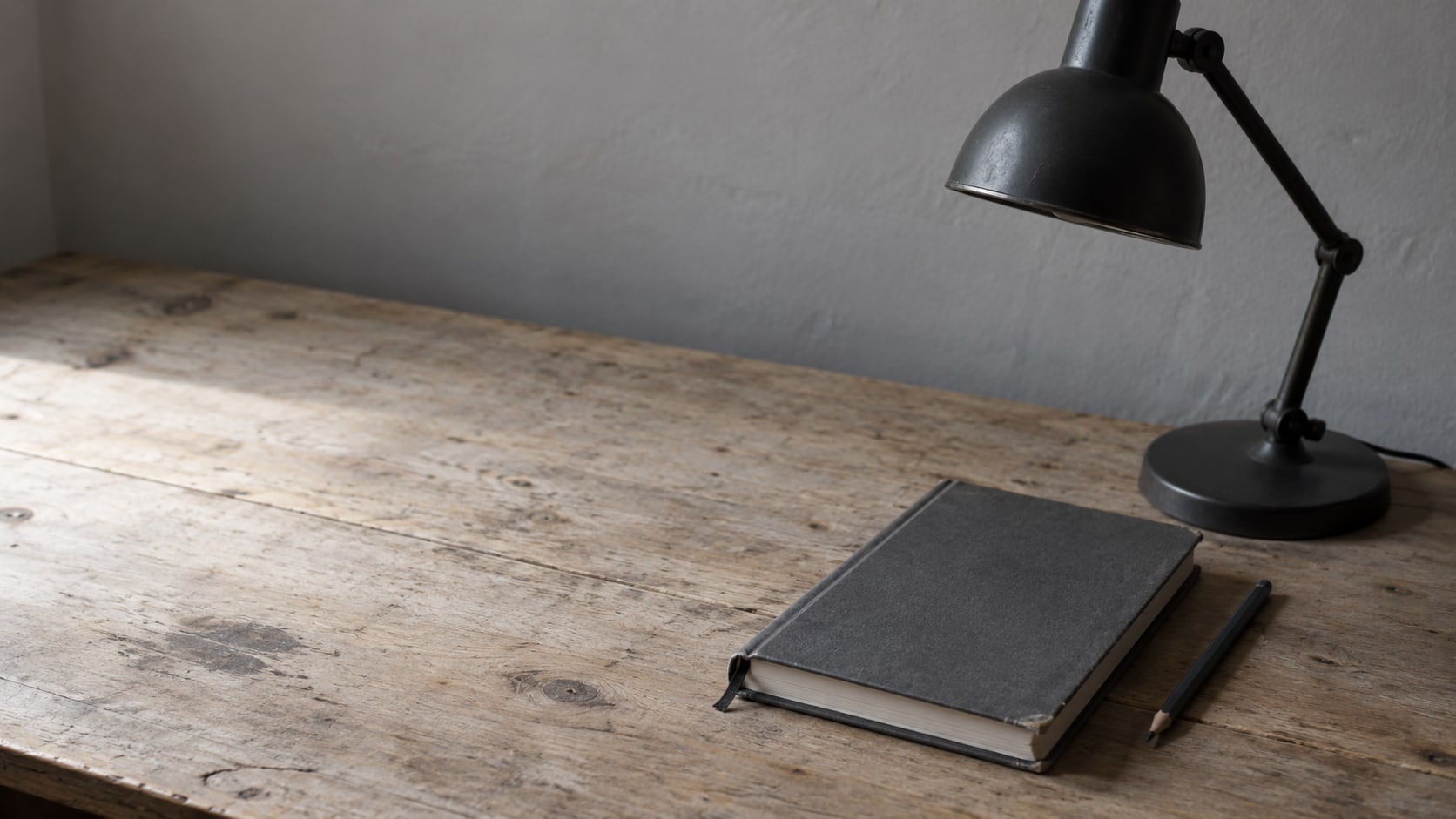 A closed appointment book and a pencil beside a switched-off desk lamp on a worn wooden desk.