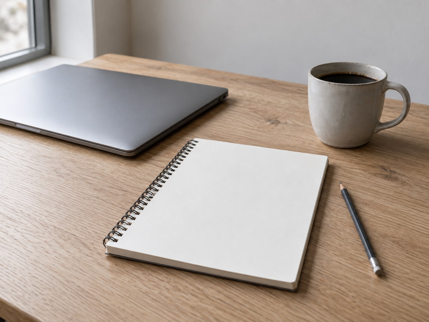 A closed laptop beside a blank notepad with a pencil and a mug of coffee on a light oak desk.