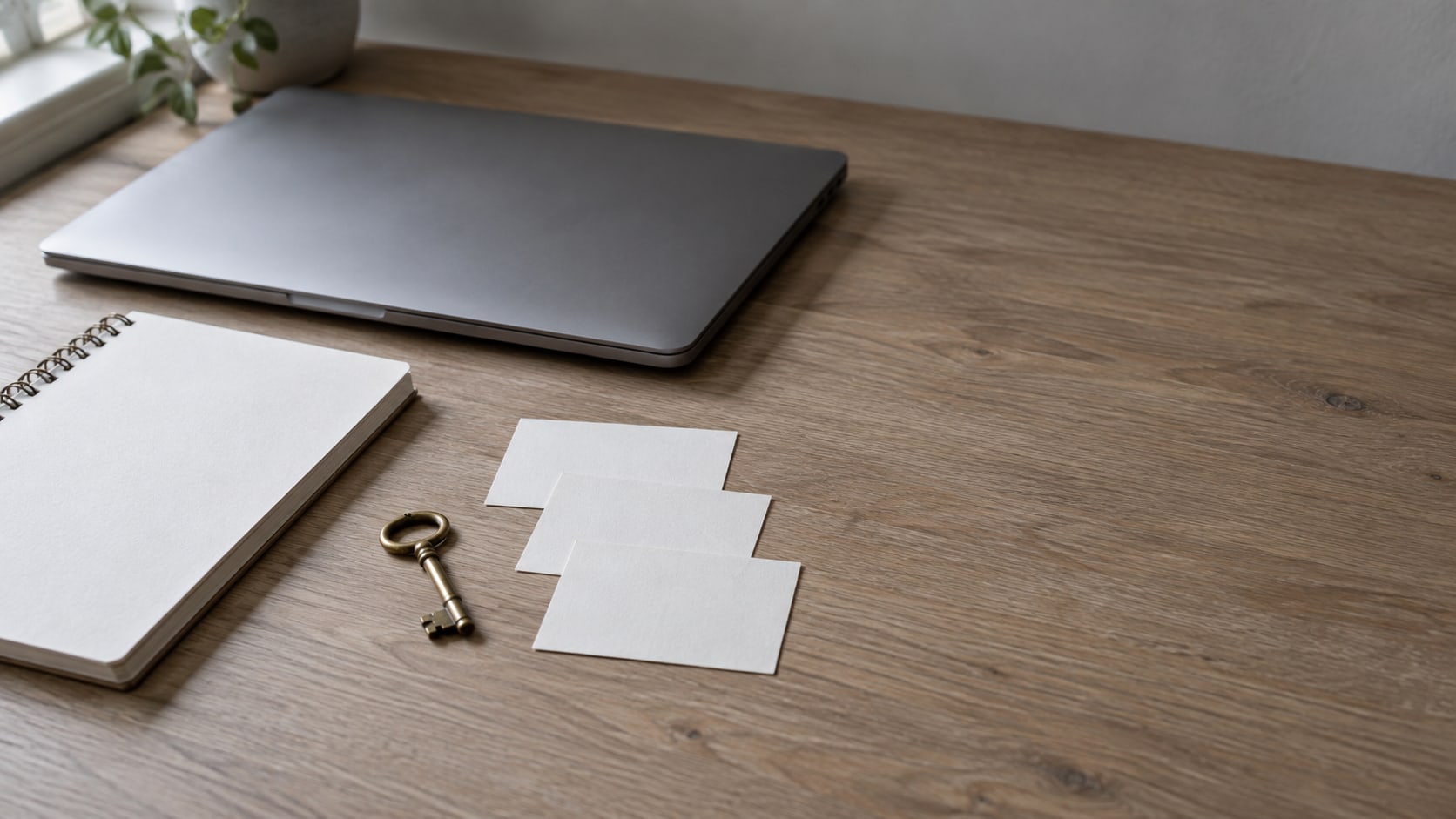 A closed laptop, a blank notepad and three plain index cards arranged beside a small brass key on a light oak desk.