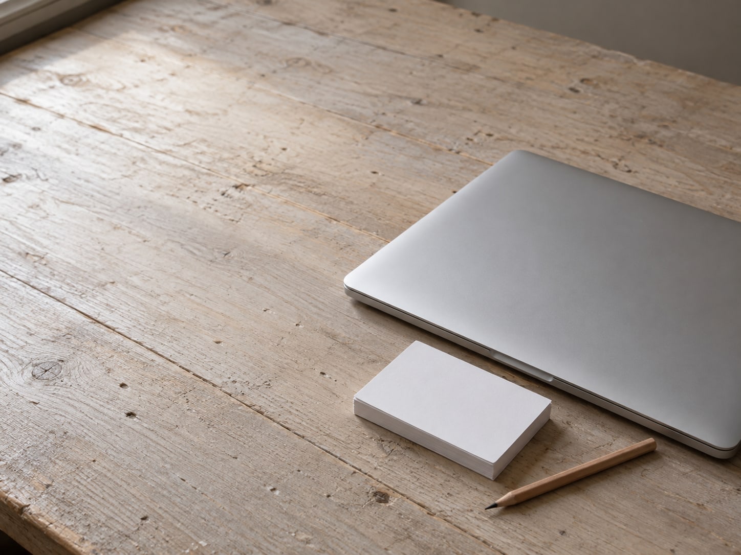 A closed silver laptop on a pale wooden desk next to a small stack of blank index cards and a pencil.
