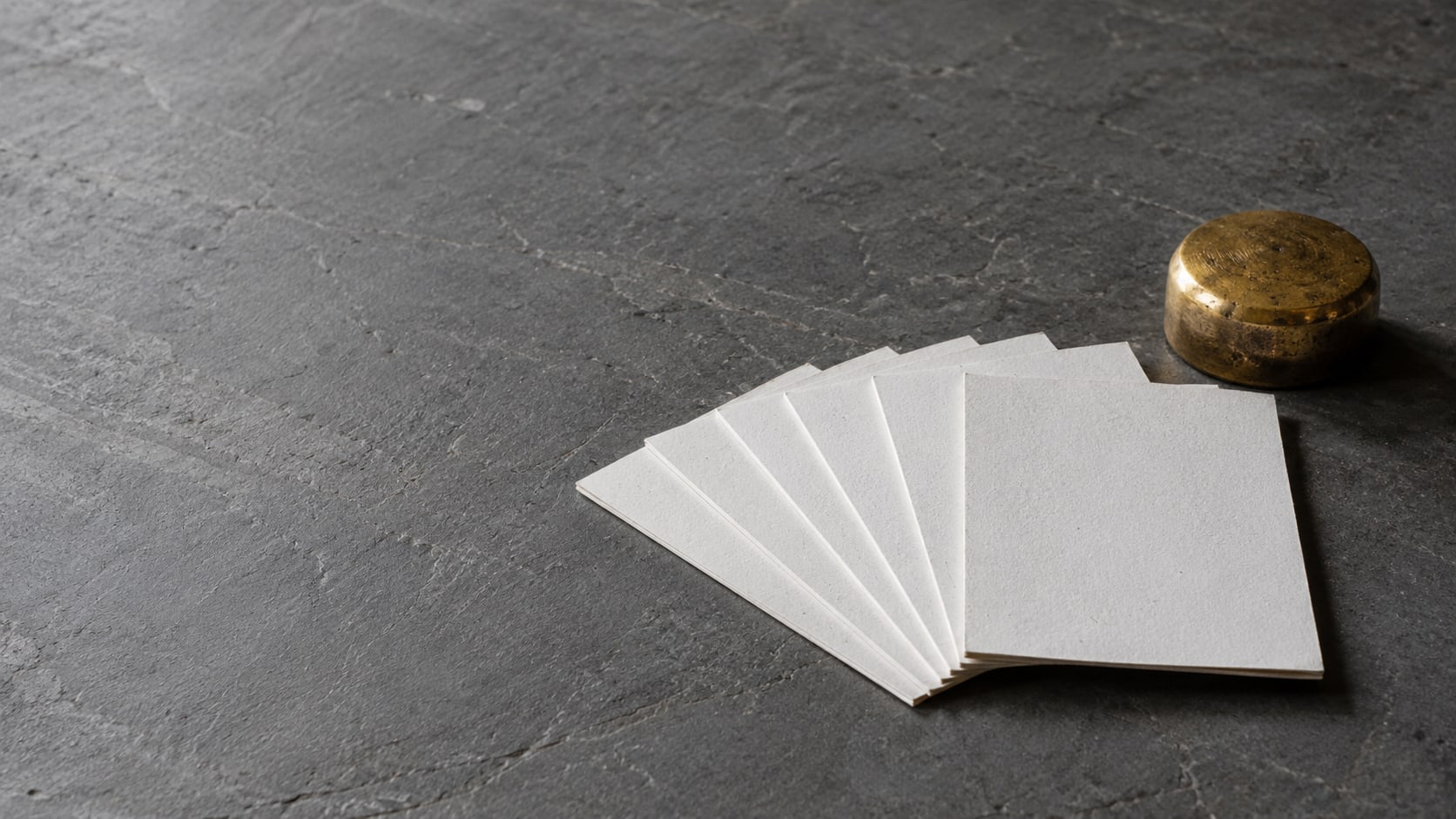 A row of identical blank white cards fanned out on a slate-gray table beside a small brass paperweight.