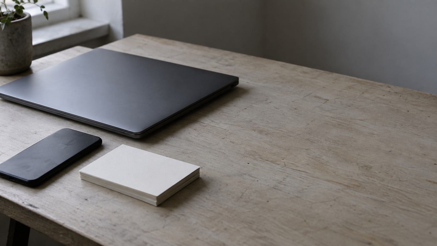 A closed laptop and a small stack of blank index cards beside a switched-off phone on a plain wooden desk.