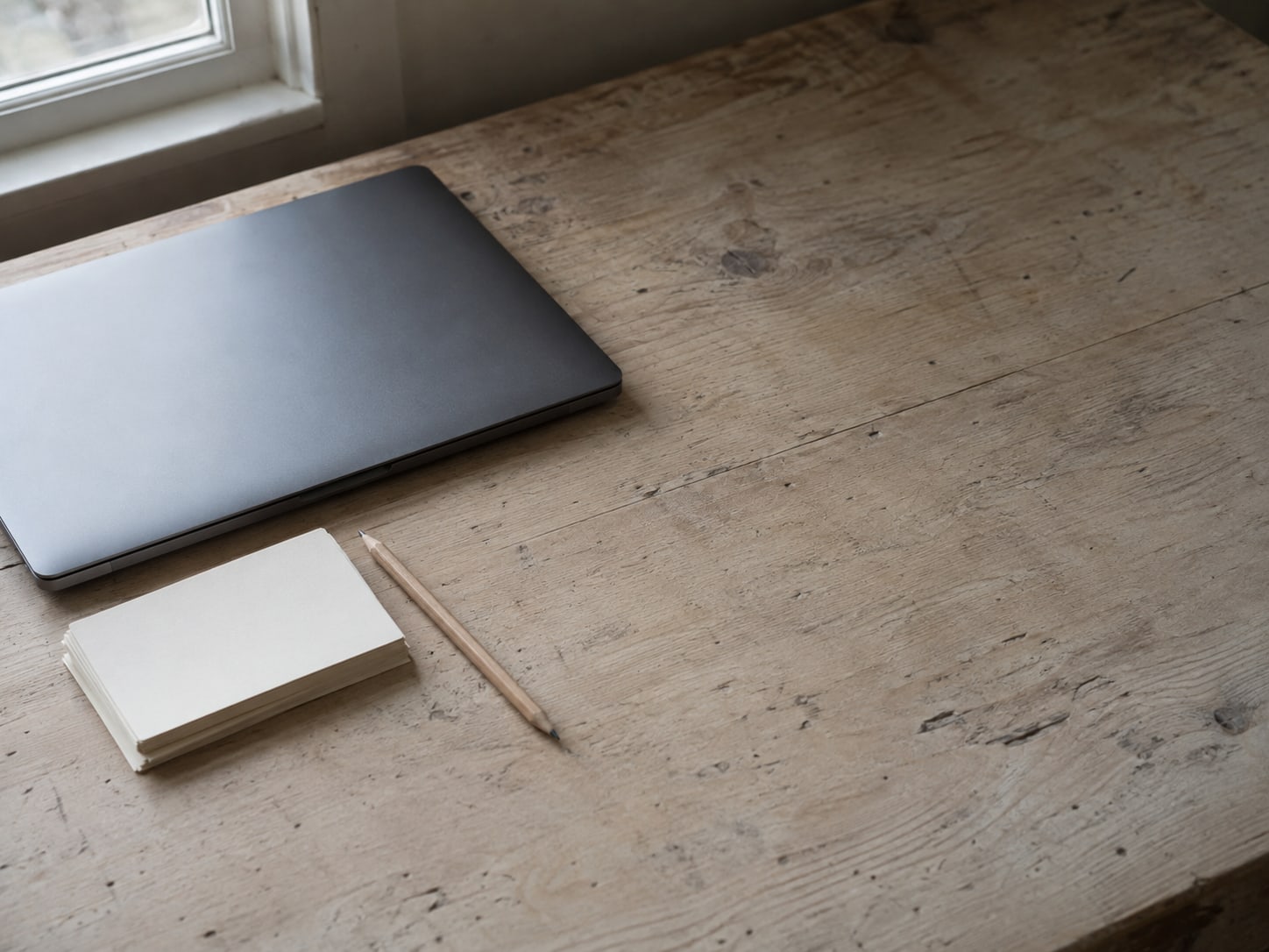 A closed laptop beside a small stack of blank index cards and a pencil on a worn wooden desk.