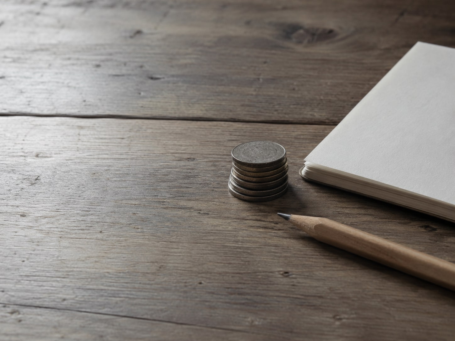 A small stack of coins beside a pencil and a blank notepad on a wooden desk.
