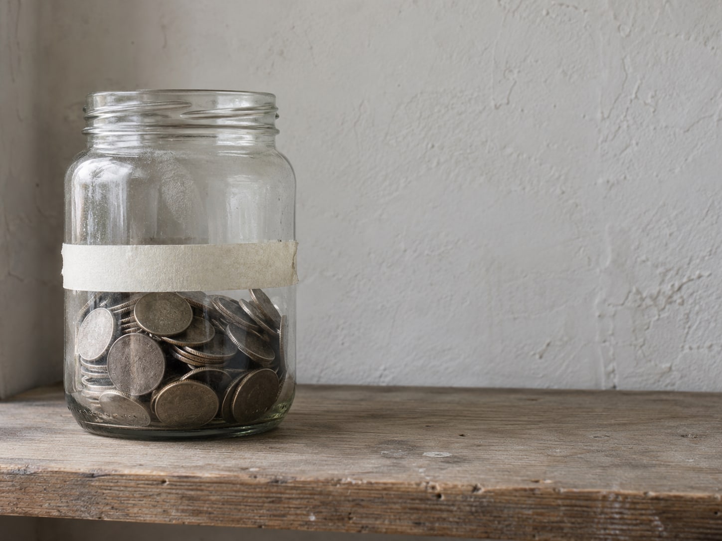 A glass jar of coins with a strip of masking tape marking a level near the middle, sitting on a wooden shelf.