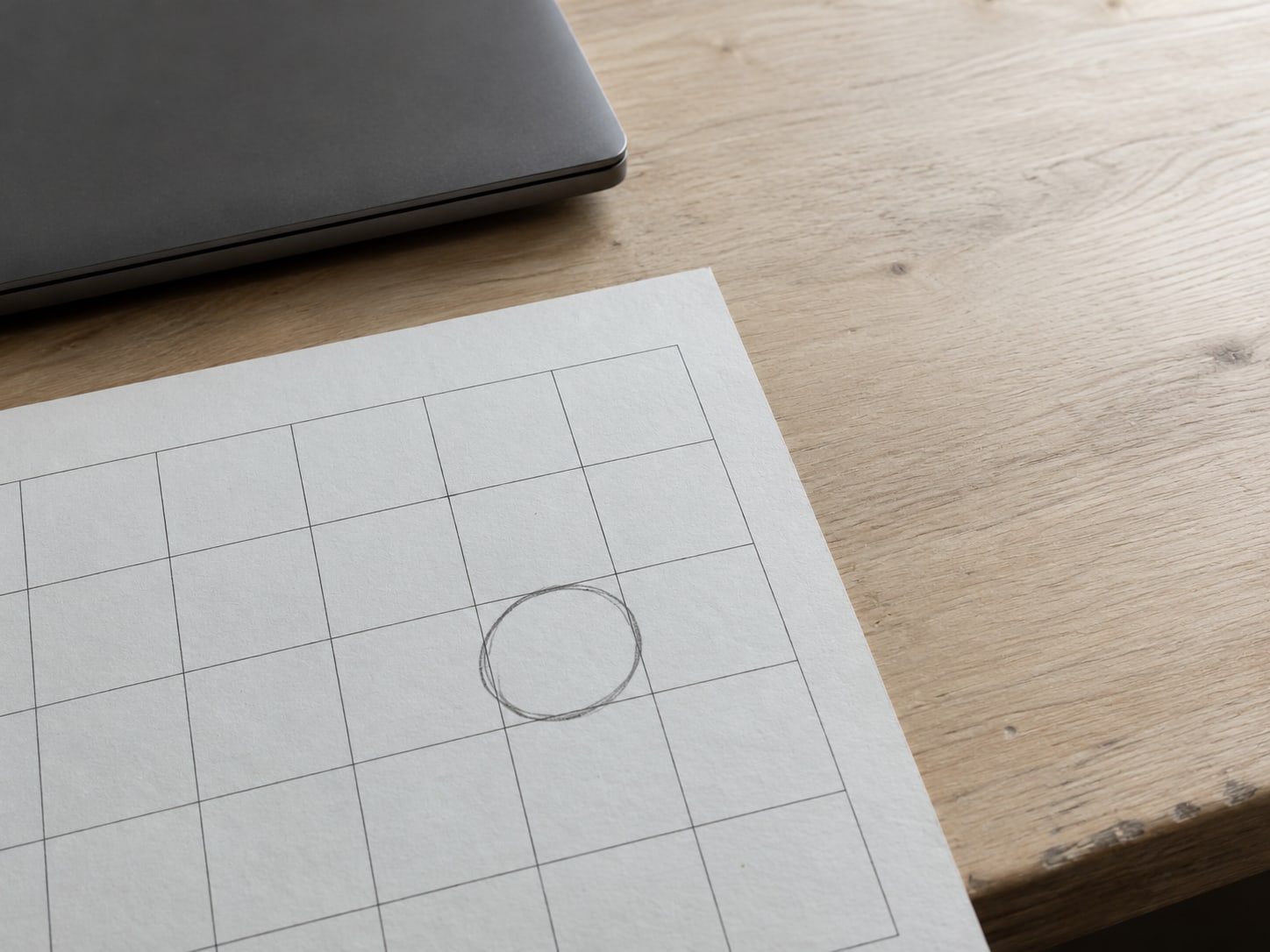 A blank paper calendar grid on a desk with one square circled in soft pencil beside a closed laptop.