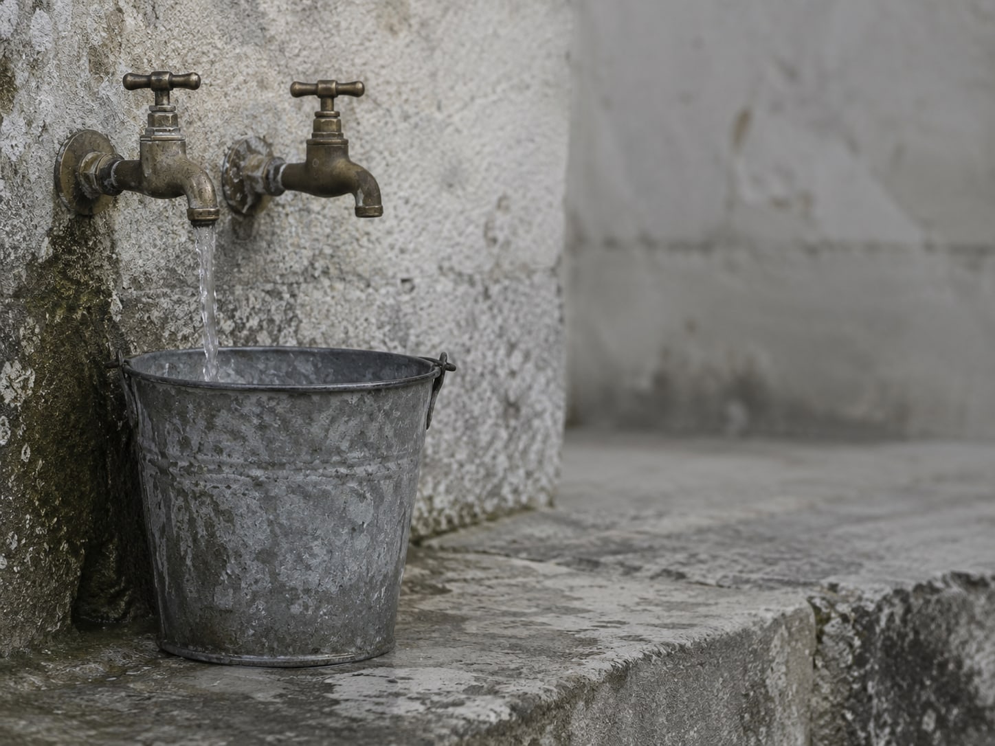 Two brass garden taps over a galvanized bucket on a stone step, one tap open a little and the other closed.