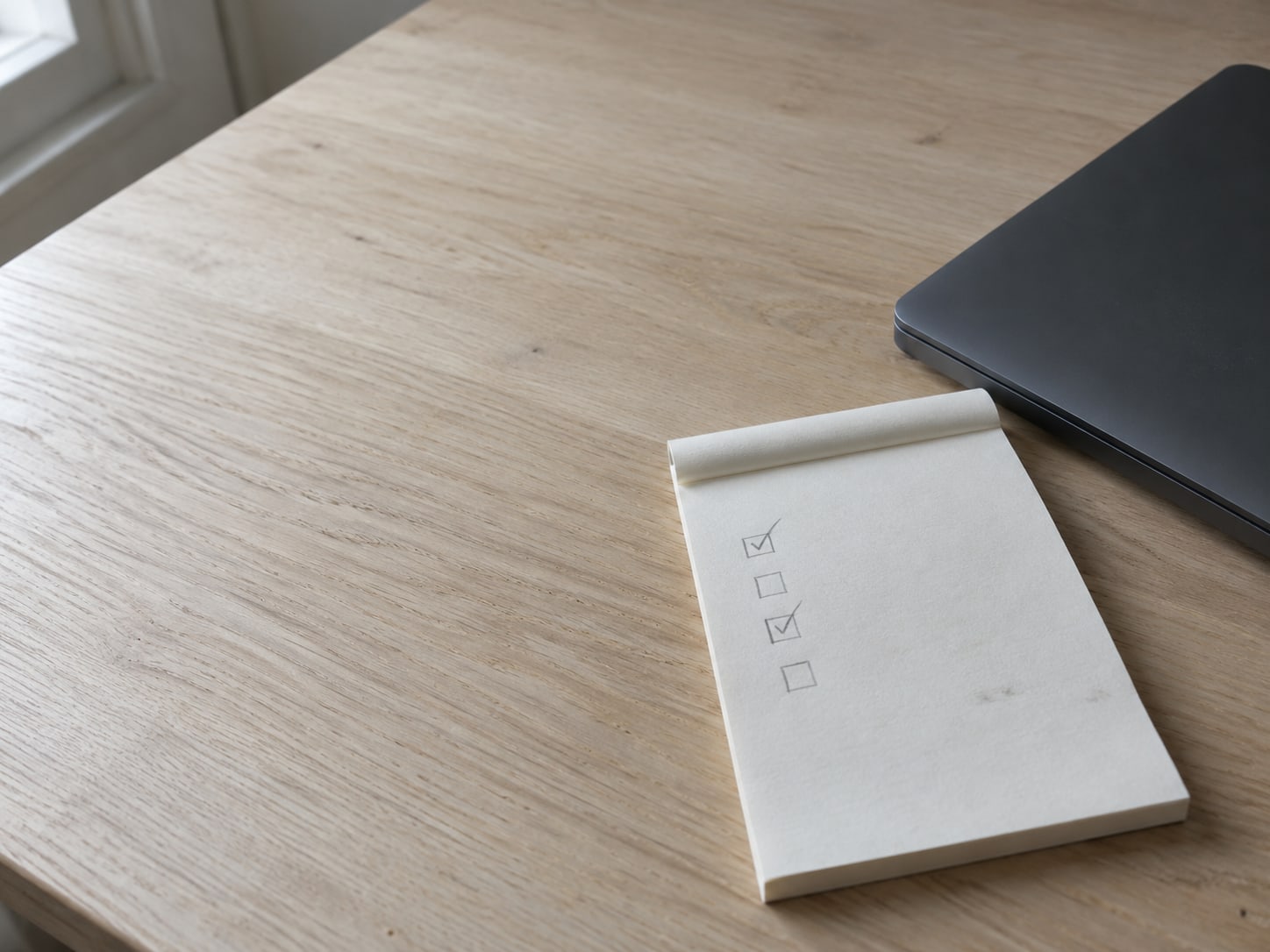 A notepad with a short pencil checklist beside a closed laptop on a clean wooden desk.