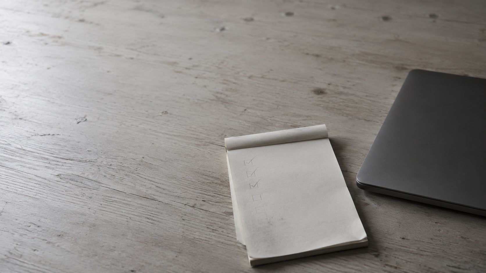 A notepad with a short pencil checklist beside a closed laptop on a clean wooden desk.
