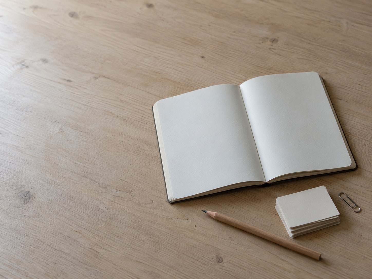 An open notebook with a pencil, a small stack of blank paper slips and a paper clip on a desk.