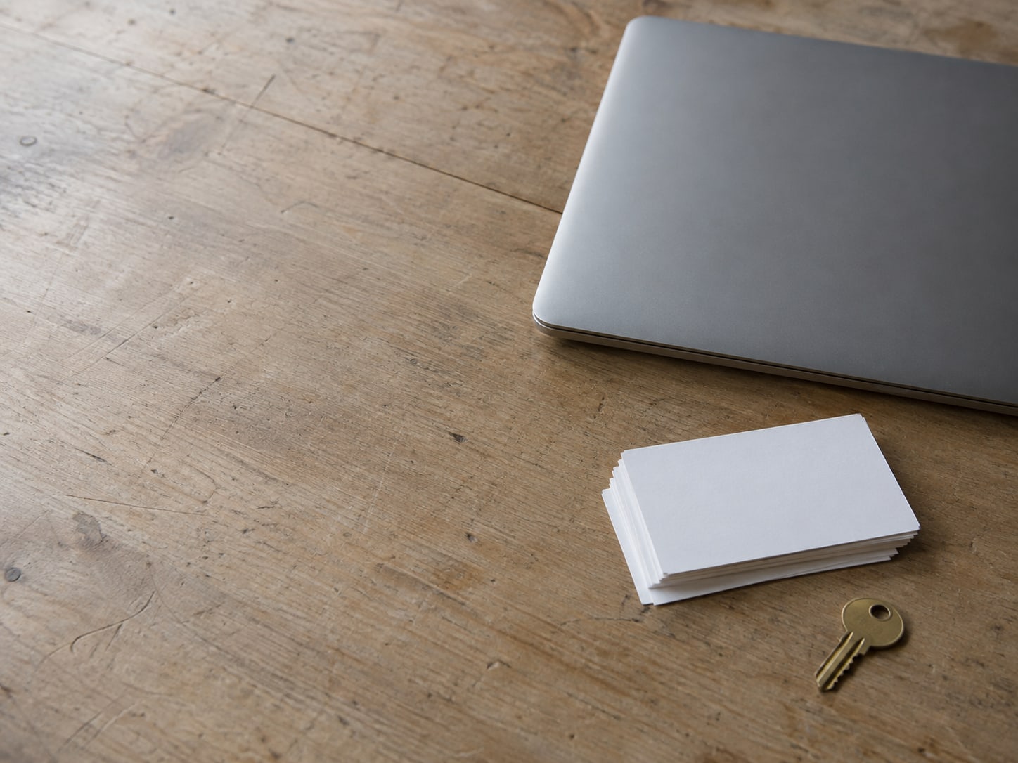 A closed laptop beside a small stack of blank index cards and a single brass key on a wooden desk.