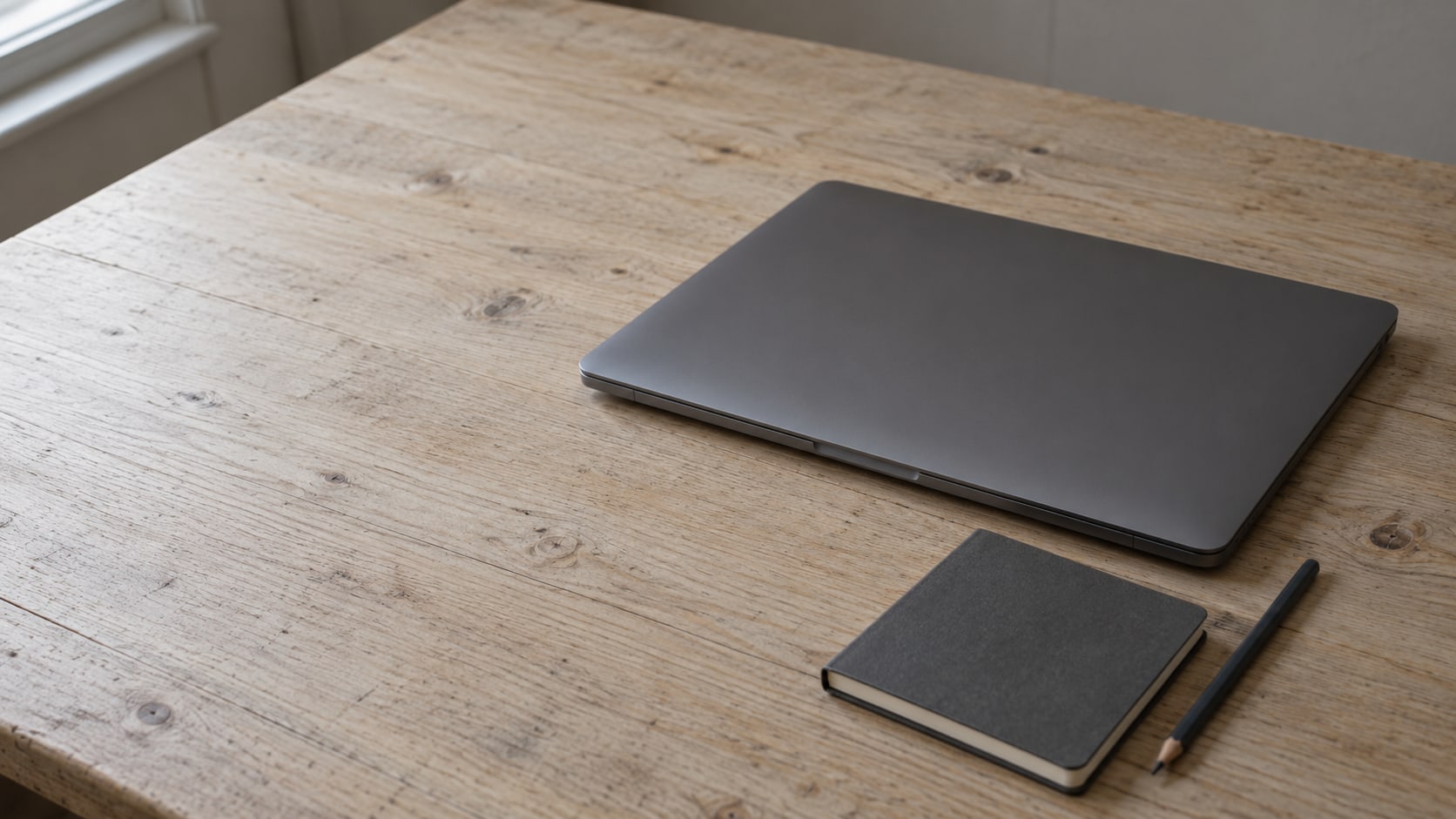 Una laptop cerrada, una libreta pequeña y un lápiz sobre un escritorio de madera limpio.